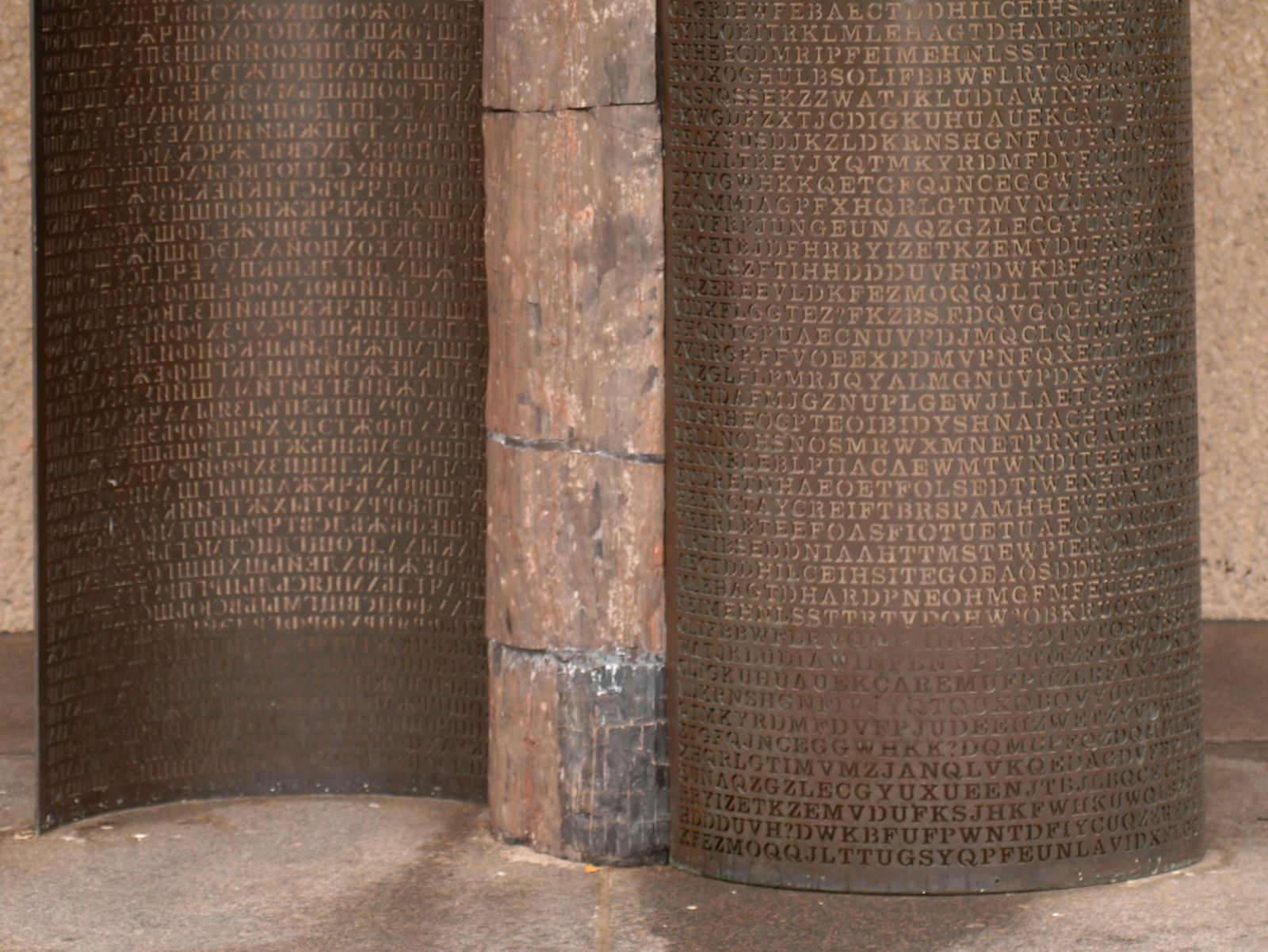 Two curved bronze panels densely engraved with capital letters, Cyrillic on the left and Latin on the right, meeting at a weathered petrified wood column. The Latin panel carries the Kryptos ciphertext.