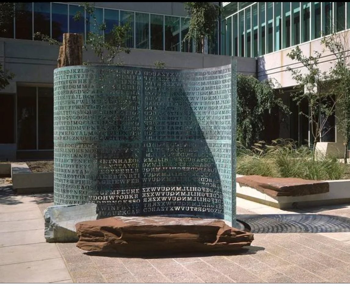 The Kryptos sculpture in the CIA courtyard: a tall S-curved copper screen, weathered to green patina, with thousands of capital letters cut clean through it. The left half carries the four encrypted messages; the right half carries a mirrored alphabet chart. A petrified tree trunk stands at the left edge and slabs of red granite lie in front.