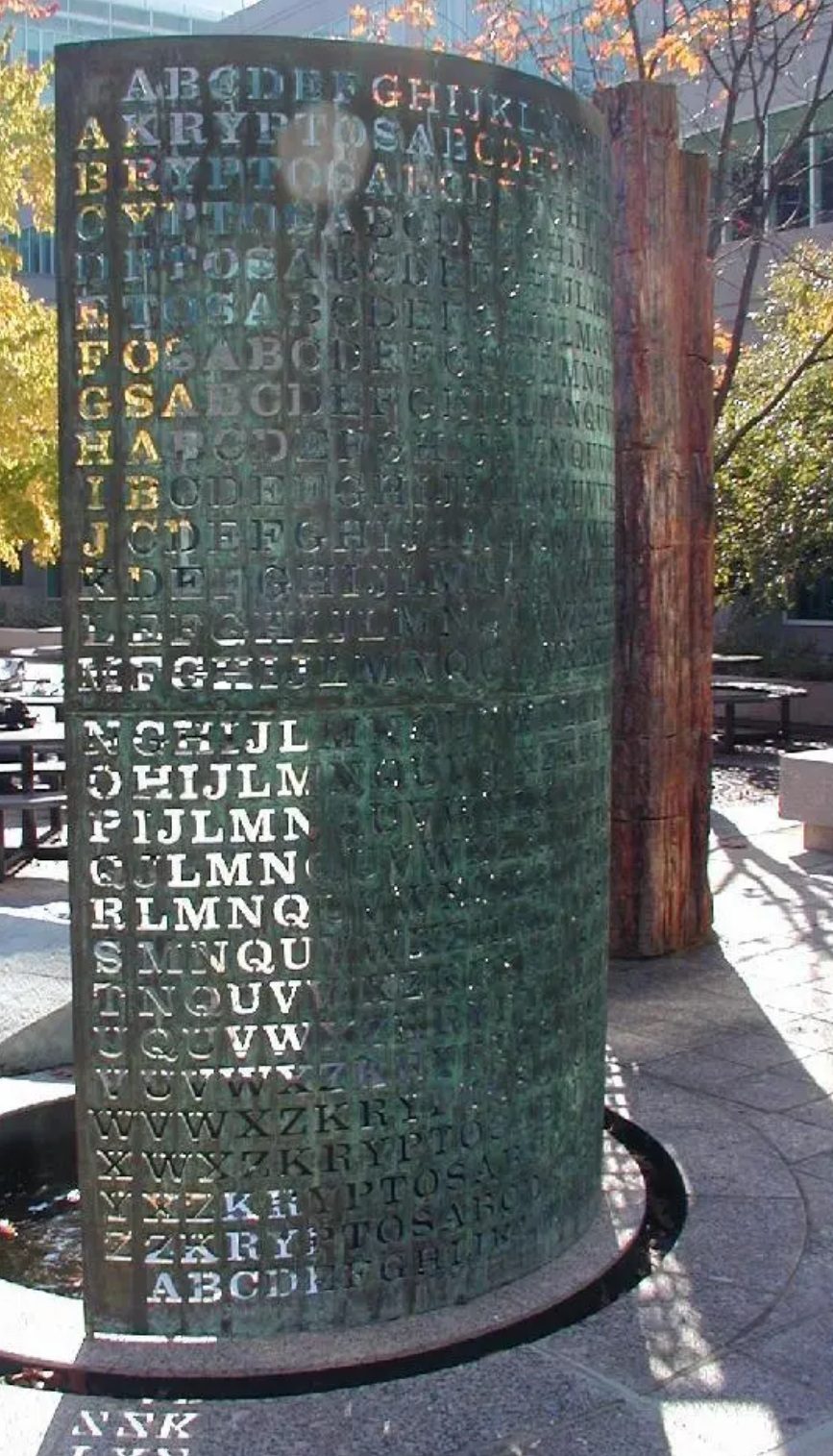 Close view of the right-hand half of the Kryptos screen, seen from behind. Twenty-six rows of capital letters are cut through the green copper. The row beginning with A reads KRYPTOSABCDEF, and each row below it begins one letter further along the same sequence.